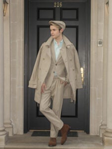 Male model wearing a classic tan trench coat and suit posing in front of a black architectural door.